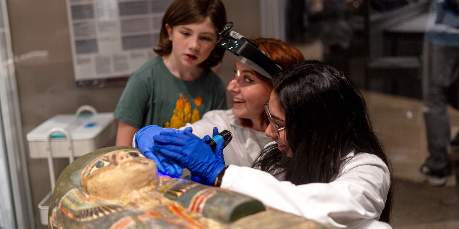 Conservators closely examining and restoring an ancient sarcophagus with blue-gloved hands using a handheld light.